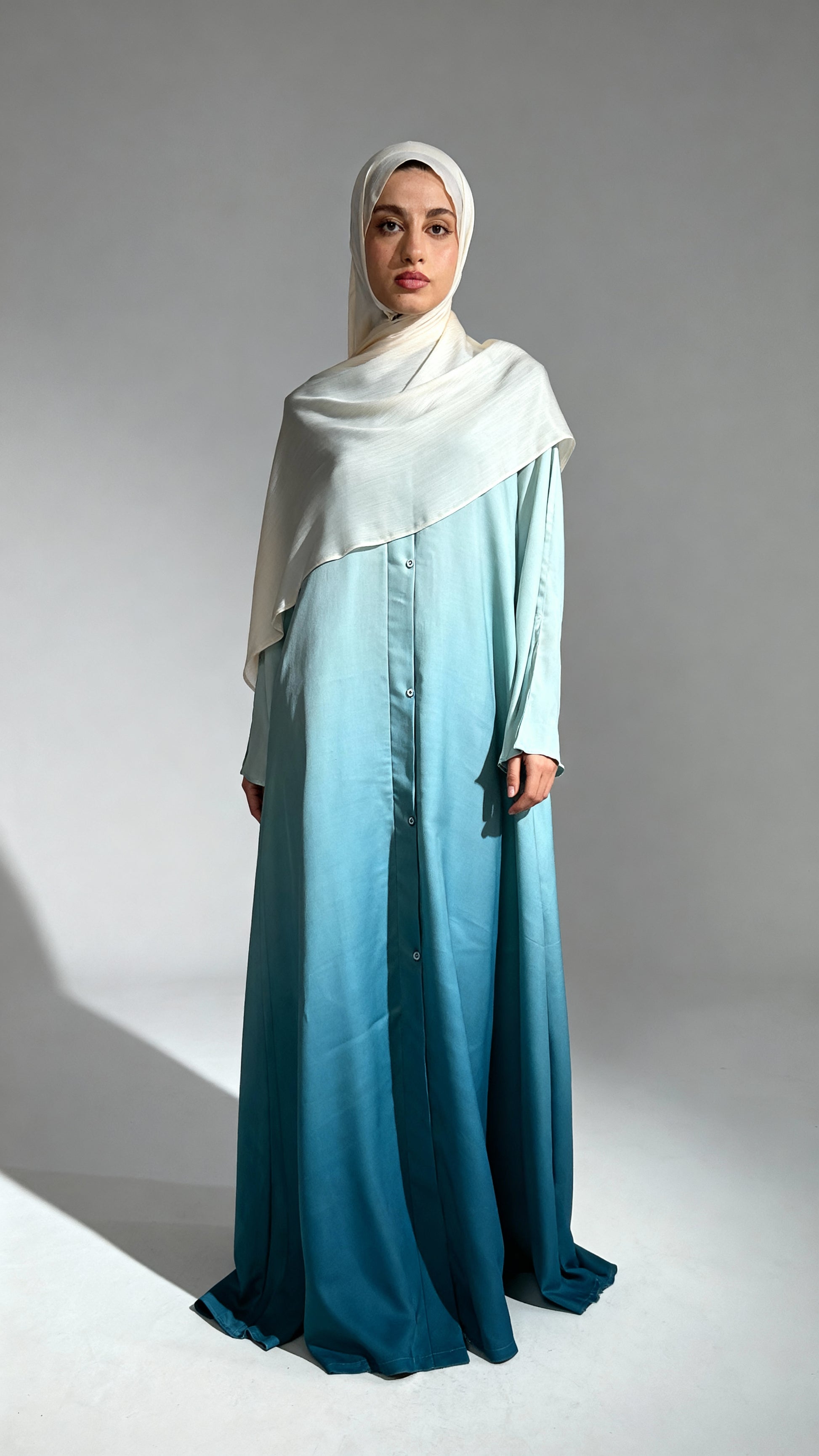 Woman wearing a light blue dress and white hijab against a gray background